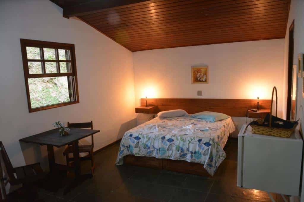 Quarto da pousada moinho azul com cama de casal, mesa e cadeiras de madeira, frigobar ao canto e janela, ilustrando post pousadas em Teresópolis.