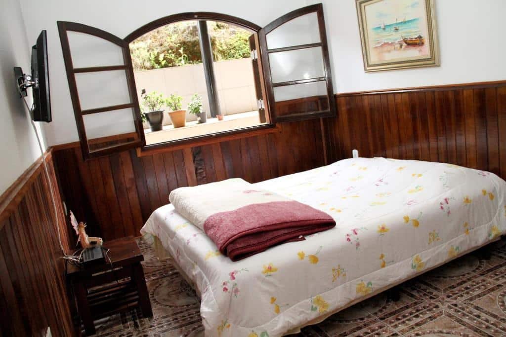 Uma cama de casal no lado direito e uma televisão no lado esquerdo. Atrás, uma janela. Quarto da Pousada Sol da Montanha.
