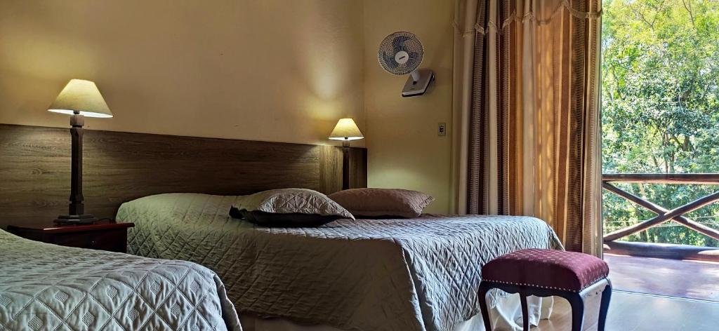 Quarto da pousada taverna do golf  em Teresópolis com duas camas, abajur, ventilador na parede e sacada com vista para as árvores, ilustrando post pousadas em Teresópolis.