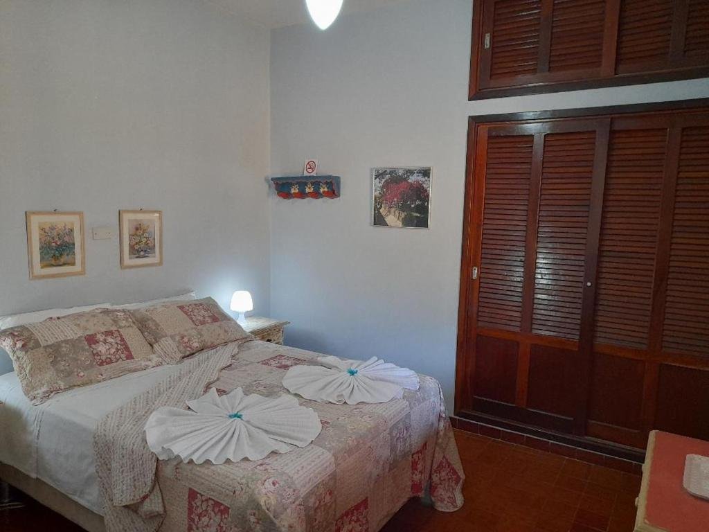 Um quarto na Pousada Suítes Águas de São Pedro, para ilustrar o post sobre pousadas em Águas de São Pedro. Há uma cama de casal e uma mesa de cabeceira com um abajur. Dois quadros estão enfeitando a parede na qual a cama está encostada. Um guarda-roupa de madeira embutido na parede está do lado direito da cama.