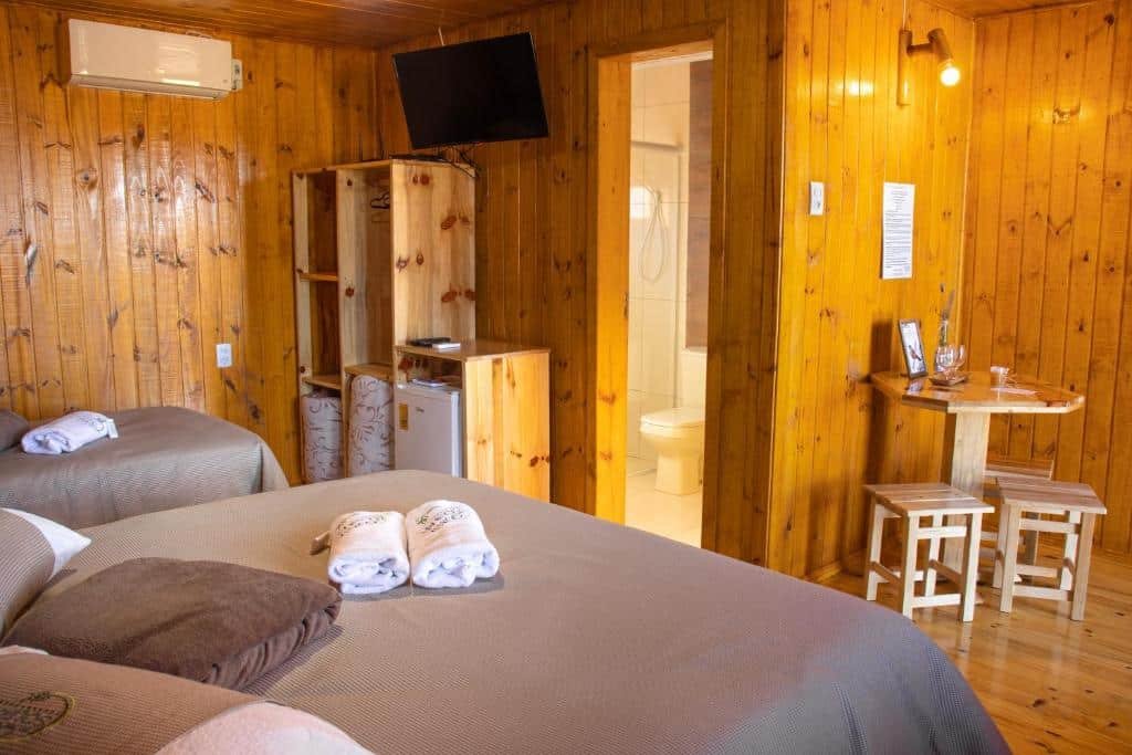 interior do chalé com uma cama de casal e uma de solteira no canto esquerdo da imagem. Em frente há um pequeno armário de madeira, sem portas, um frigobar, uma porta aberta com acesso ao banheiro e uma pequena mesa redonda de madeira com três banquinhos envolta.