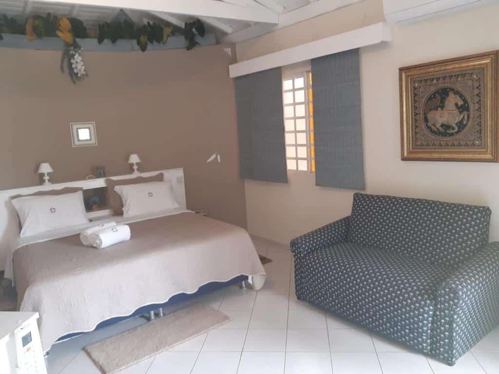 Quarto em Recanto Sereníssima, representando o post de pousadas em Águas de São Pedro. A decoração do ambiente é elegante, possui uma cama de casal box com pequenos abajures em cima de sua cabeceira, e um mini sofá pontilhado está encostado na parede em frente a cama. A janela da suíte está do lado direito da cama.