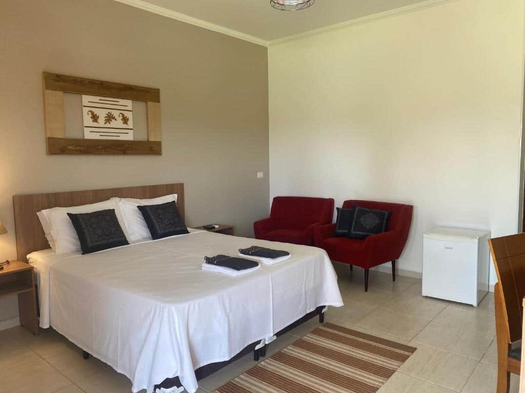Imagem de um quarto da Pousada San Pietro, representado o post sobre pousadas em Águas de São Pedro. O quarto é espaçoso, possui uma cama box de casal, do seu lado temos visão de duas poltronas vermelhas e um frigobar.