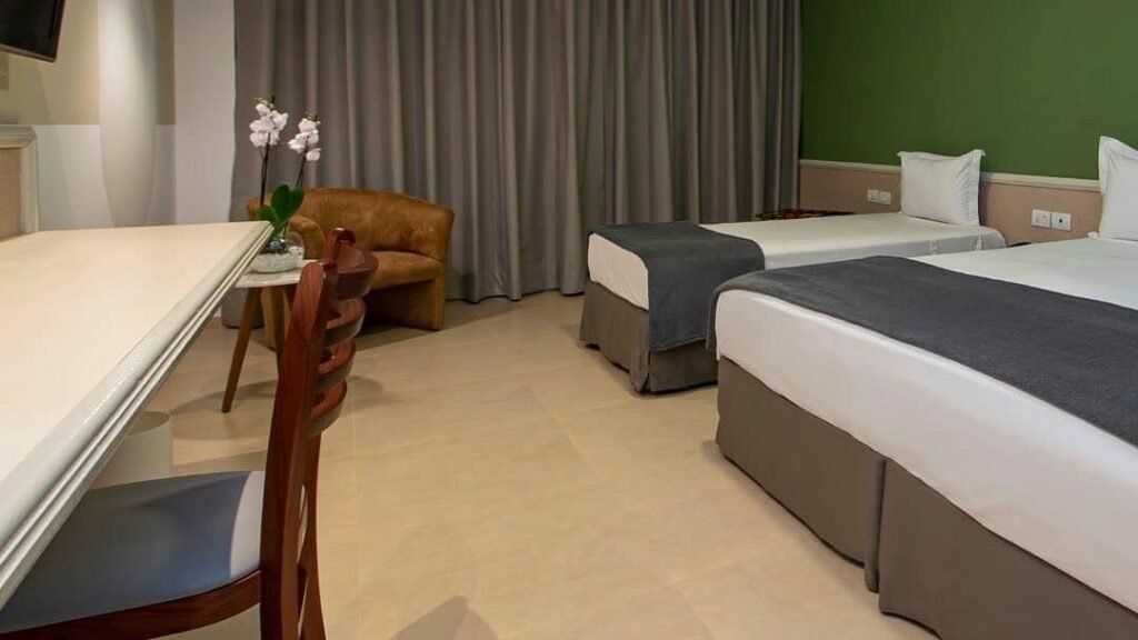 Quarto no Villagio Embu. Uma cama de casal e uma cama de solteiro no canto direito. No canto esquerdo uma mesa de trabalho e uma televisão. Atrás uma mesinha com um arranjo de flor e uma poltrona.