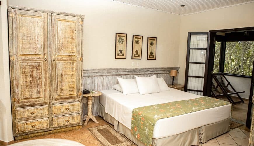 Quarto no . Um guarda-roupa no canto esquerdo, ao lado uma mesinha de apoio e uma cama de casal. No canto direito uma porta para a sacada com duas cadeiras de madeira.
