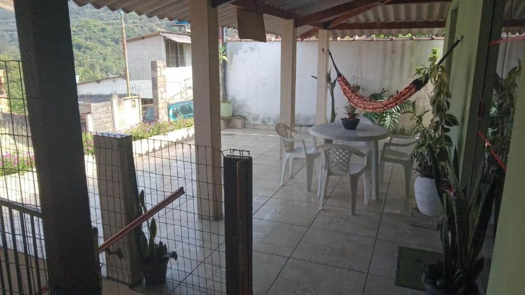 Área externa do airbnb Cantinho da Serra. No lado direito é possível ver a porta de vidro que dá acesso a área externa da casa. Em uma área coberta, no centro da imagem, há uma mesa de plástico com cadeiras e uma rede de descanso. Há quatro pilastras no lado esquerdo da imagem.
