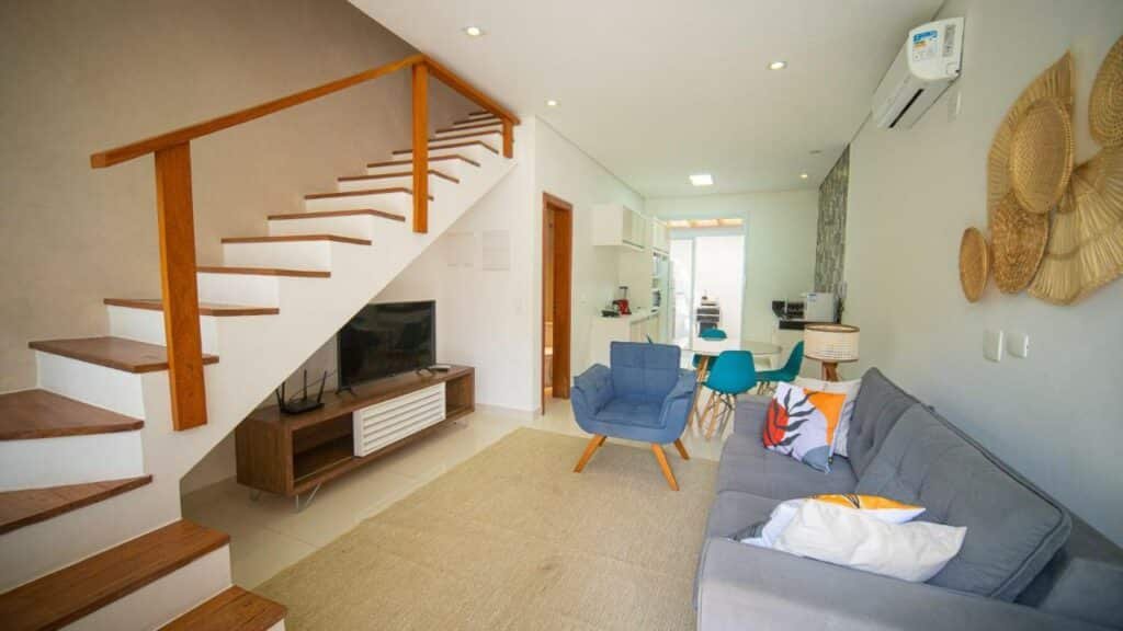 Sala de estar do Residencial Las Salinas. Ao fundo está a cozinha e uma porta que leva para o exterior, já em primeiro plano, na sala, um sofá está colado na parede direita e de frente para uma tv, que está debaixo da escada. Entre eles há uma poltrona e um tapete.