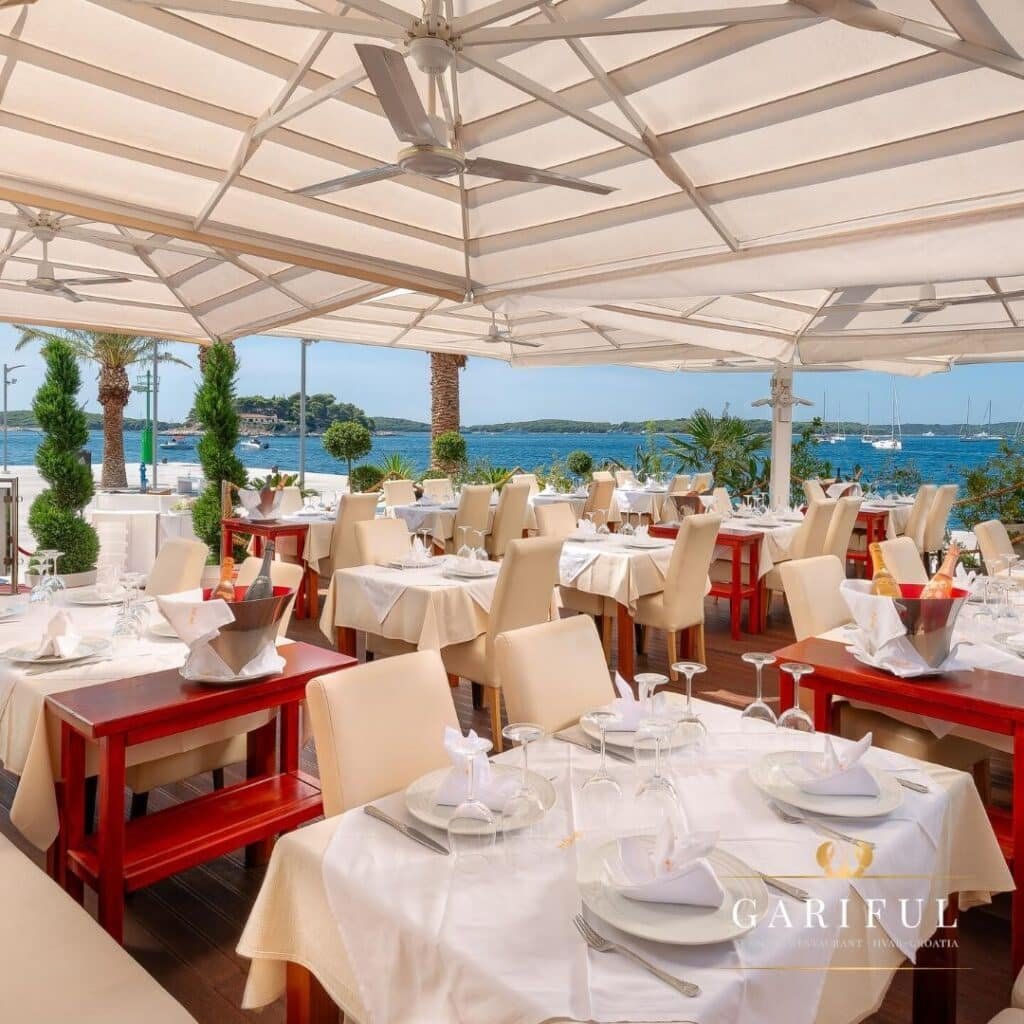 Restaurante Gariful em Hvar ao ar livre com mesas para refeições, sob marquises brancas, com vista para uma orla pitoresca com palmeiras e barcos.