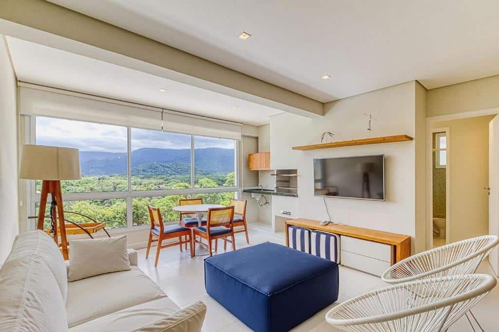 Sala de estar do D21 - Conforto junto a natureza - Praia de Camburyzinho durante o dia com sofá do lado esquerdo da imagem e do lado direito duas cadeiras ao fundo uma mesa com cadeiras redondas com quatro cadeiras. Representa airbnb em São Sebastião.