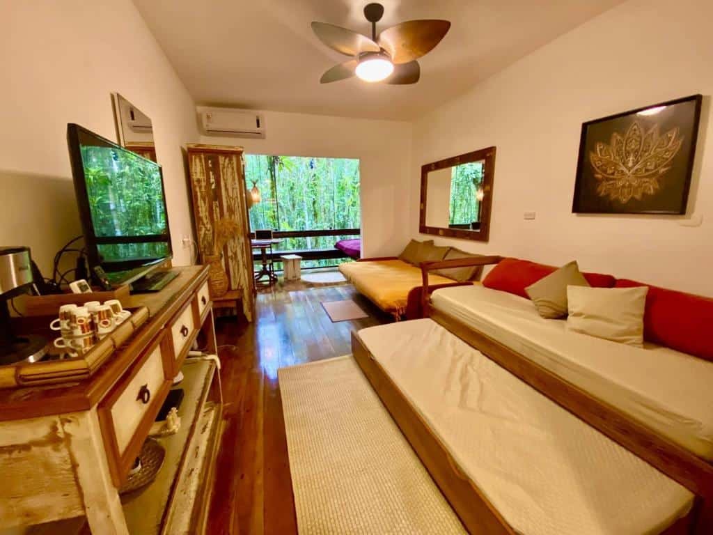 Sala de estar do Maresias, Chalé H29 em condomínio com sofá-cama a frente do lado direito da imagem e em frente ao sofá-cama uma cômoda com TV ao fundo outro sofá-cama do lado direito.