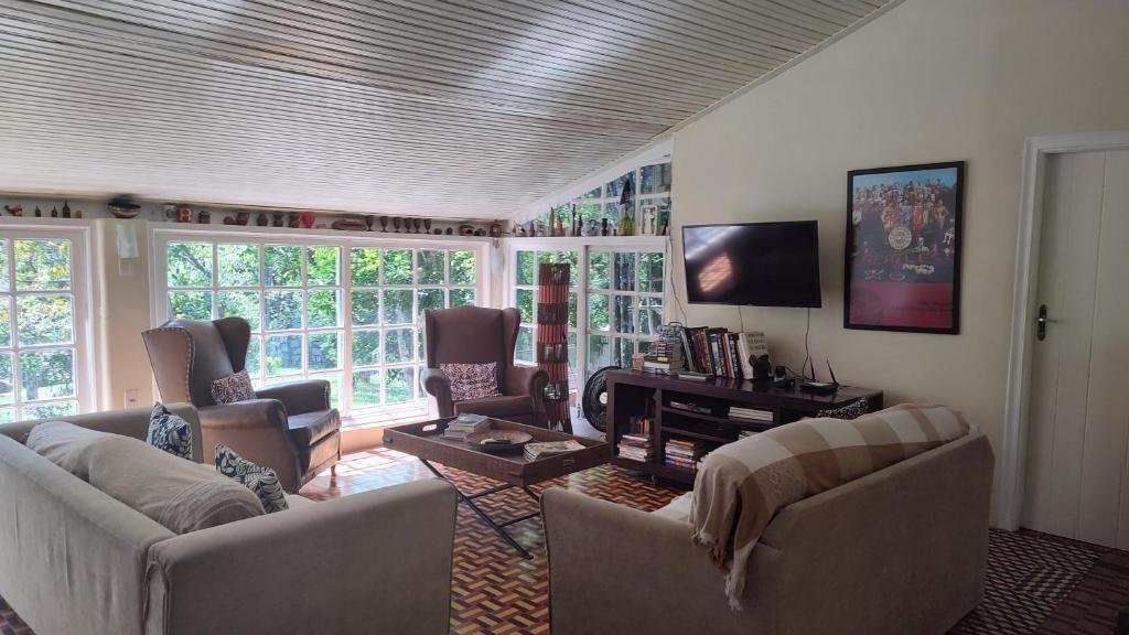 Sala de estar do airbnb Rancho São Carlos. No canto direito é possível ver um pedaço de uma porta e no lado esquerdo há um rack com livros e uma TV presa a parede. No lado esquerdo da sala a parede é composta por três janelas bem grandes. Ainda no canto esquerdo há duas poltronas grandes. No centro da sala há uma mesa de centro e no lado direito há dois sofás. 