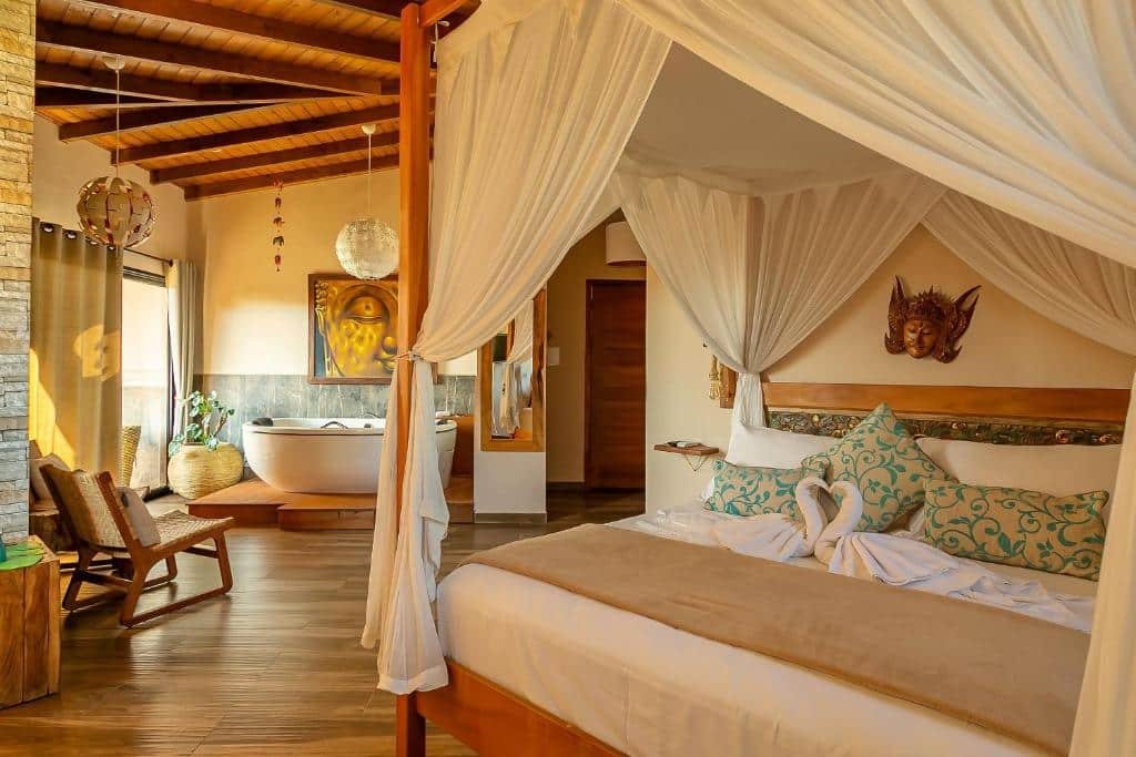 Quarto do hotel Shiva Boutique Hotel - Praia do Rosa. Uma cama de casal com dossel está do  lado direito. Ao fundo, no lado esquerdo, há uma cadeira e uma banheira encostada na parede, abaixo de um quadro e ao lado de uma porta transparente com cortinas e um vaso de planta. No meio da imagem, entre a banheira e a cama, é possível ver um espelho na parede e uma porta fechada ao fundo. 