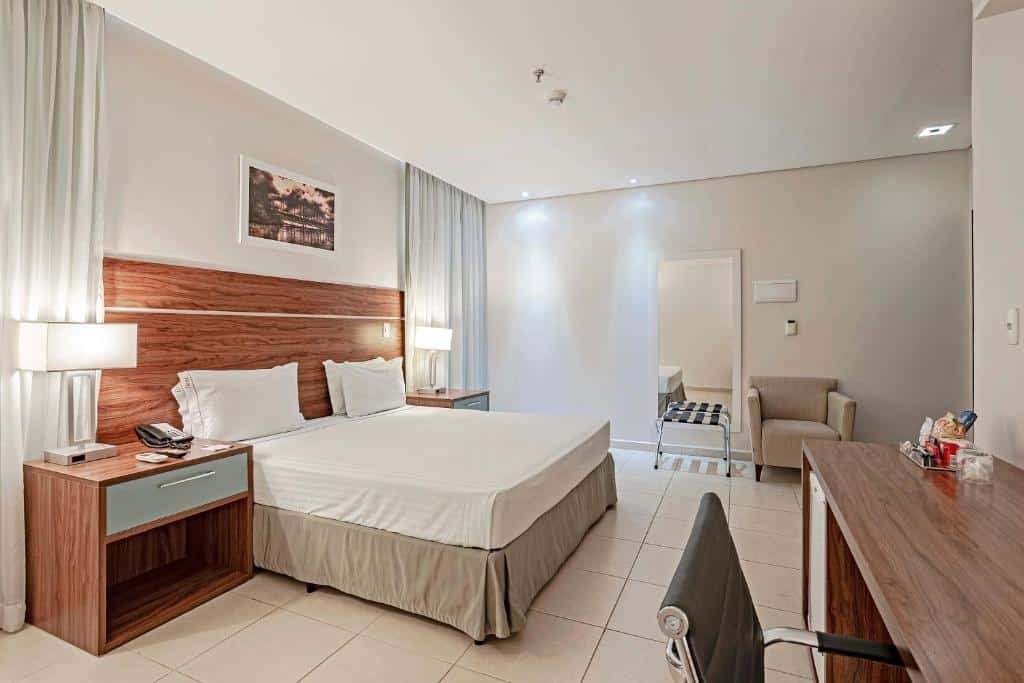 Quarto do Slaviero Porto Velho. Uma mesa de trabalho com frigobar do lado direito, do lado esquerdo uma cama de casal com uma cômoda e um abajur de cada lado, no fundo uma poltrona e um espelho. Foto para ilustrar post sobre hotéis em Porto Velho.