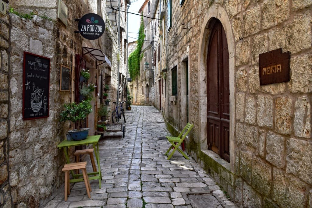 Beco estreito de paralelepípedos com um pequeno café ao ar livre em Hvar (Stari Grad), há cadeiras verdes, plantas em vasos e edifícios de pedra em ambos os lados.
