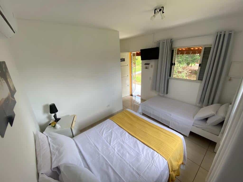 Foto do quarto na pousada Família Busca-Pé, que representa o post sobre pousadas em Águas de São Pedro. A suíte possui uma cama de casal, e ao seu lado há um frigobar. Na frente dessa cama há outra de solteiro. Uma TV está presa na parede, em direção à cama de casal. 