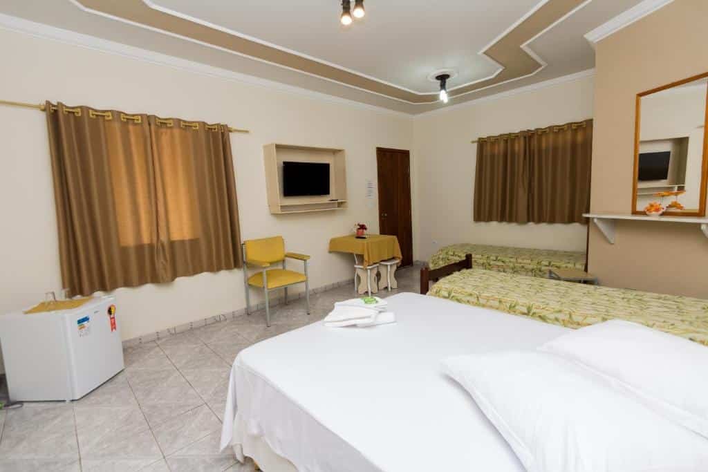 Foto de um quarto na Pousada Portal, em São Pedro, representando o post sobre pousadas em Águas de São Pedro. O quarto possui três camas, uma sendo de casal. Uma das camas de solteiro está de frente verticalmente para a porta do quarto. Mais ao fundo, as outras camas restantes estão uma do lado da outra. Uma TV está fixa na parede em frente as camas. Um frigobar está presente no canto do quarto. E uma cadeira e mesa também estão no ambiente, próximas à televisão.