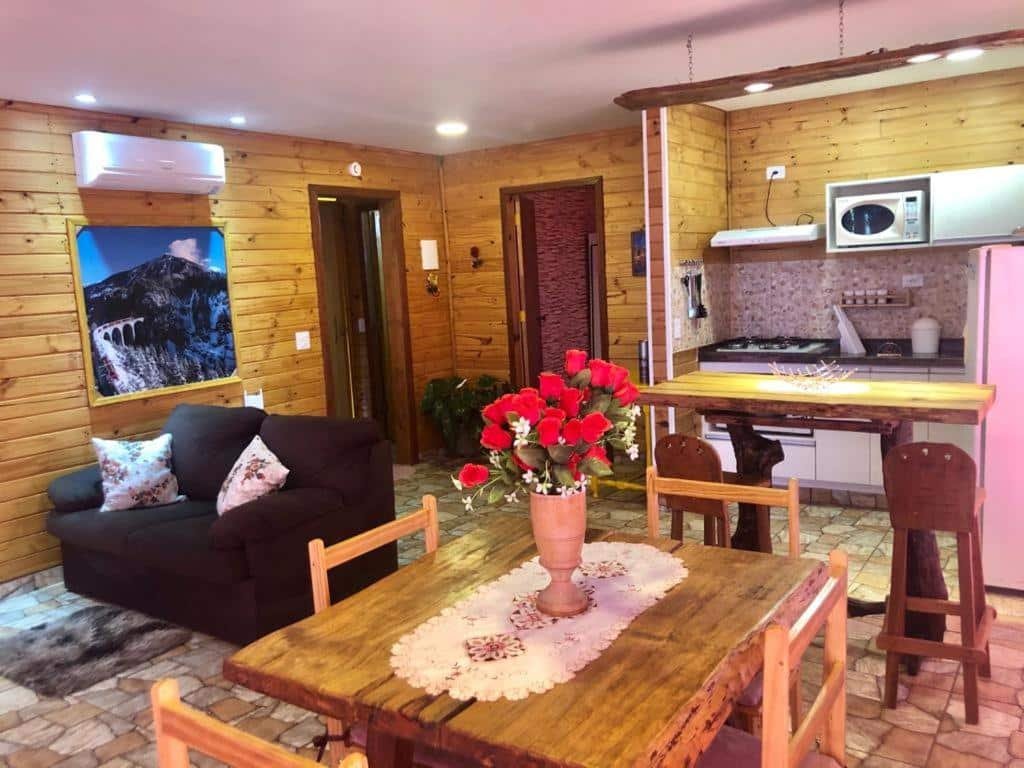 Foto  do interior de uma suíte no Swiss Chalet, para ilustrar o post sobre Águas de São Pedro. Há um sofá preto com almofadas, uma mesa de madeira com quatro cadeiras, e, mais para o fundo, outra bancada de madeira com duas cadeiras e uma cozinha equipada com fogão, geladeira, micro-ondas e armários.