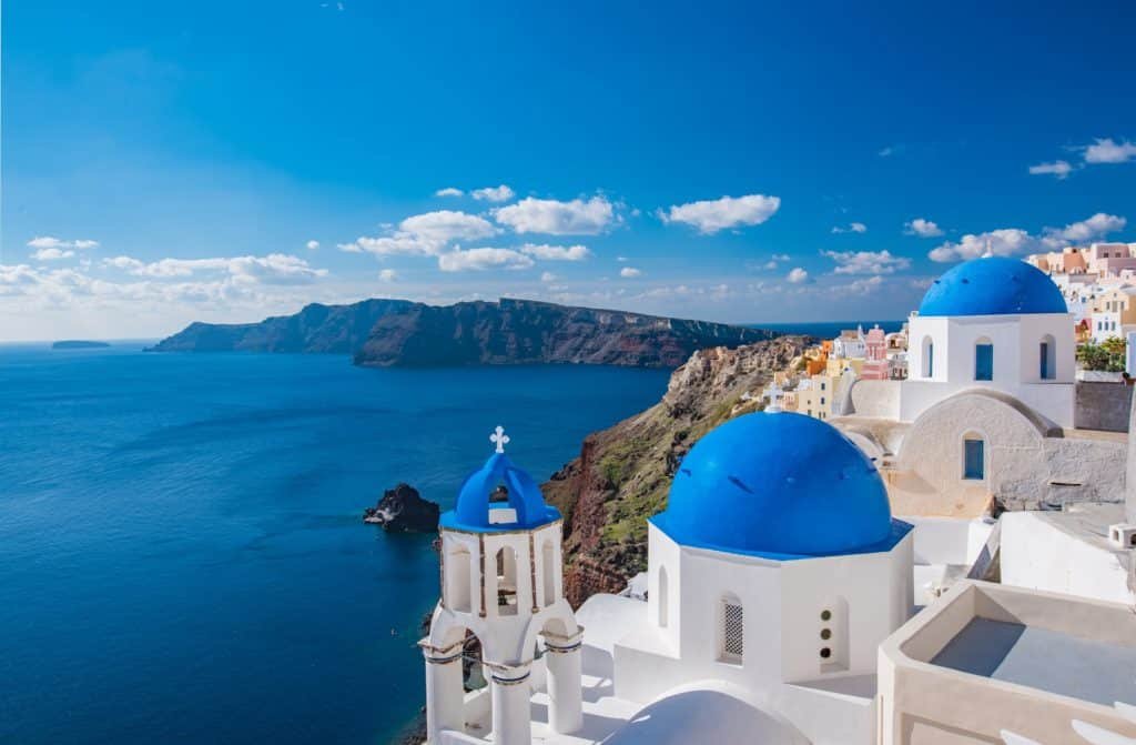 Vista de uma vila costeira em Santorini com edifícios brancos e cúpulas azuis com vista para o mar, tendo como pano de fundo colinas e céu azul.