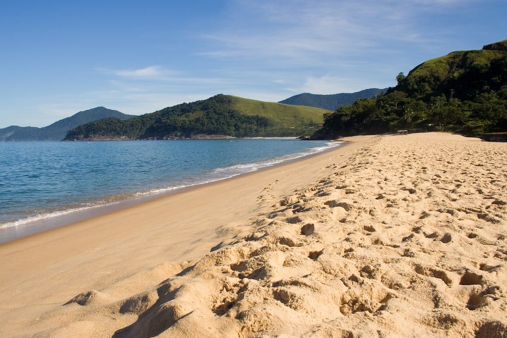 Toque-Toque Pequeno com ondas suaves e céu azul claro; colinas cobertas de verde ao longe estão sob um clima ensolarado.