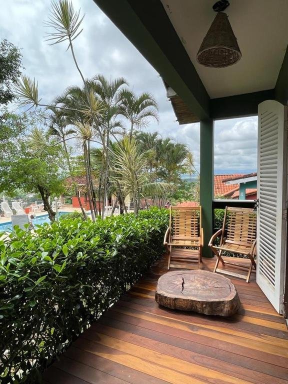 Varanda do airbnb Chale linda vista Penedo. No lado direito é possível ver a porta da varanda aberta. Na varanda há duas cadeiras e um tronco de árvore como mesa de centro. A varanda é rodeada por um grande arbusto. No fundo da imagem é possível ver diversas árvores e uma piscina.