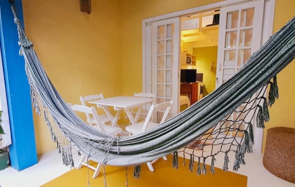 Varanda do Vilarejo Chalé. Ao fundo está a porta para a sala. Uma mesinha de quatro lugares está perto de uma rede.